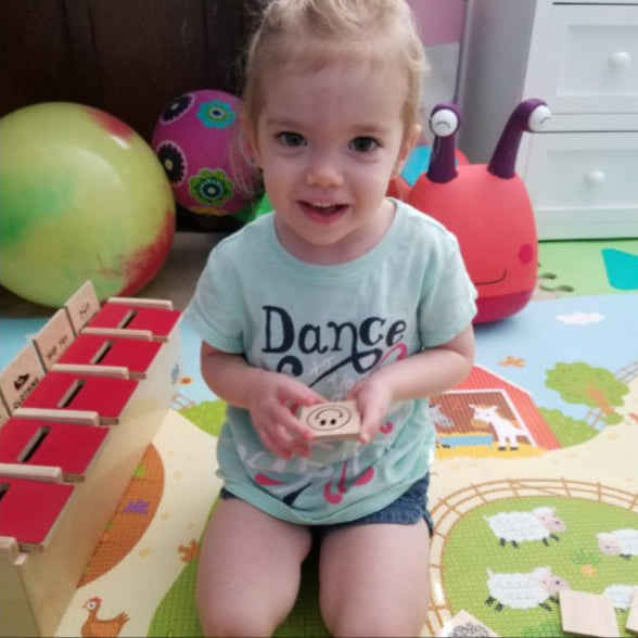 Toddler smiling while holding a wooden card with a pig illustration, sitting on a colorful farm-themed play mat.