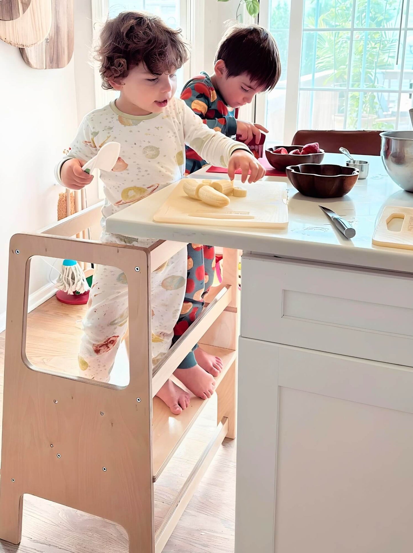 two children in learning tower working in kitchen cutting bananas with knife