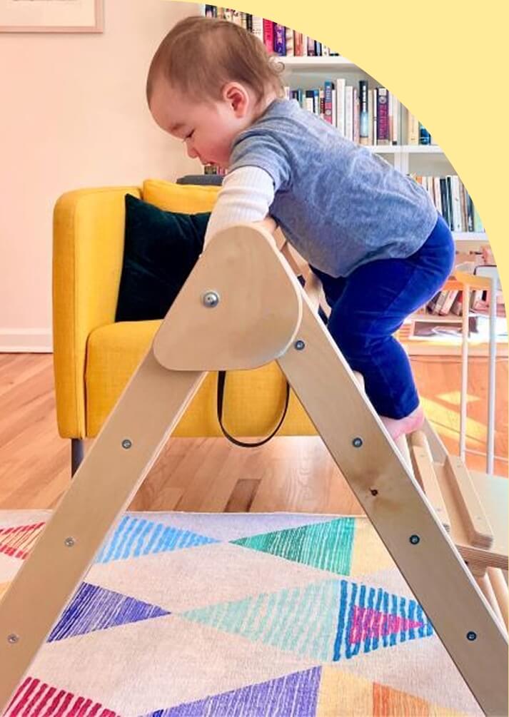 Child climbing a wooden pikler triangle in a room with books and a yellow chair.