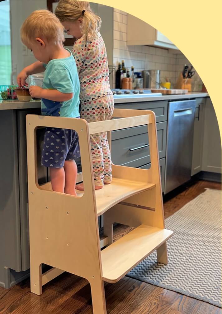 Two children using a wooden step stool in a kitchen.