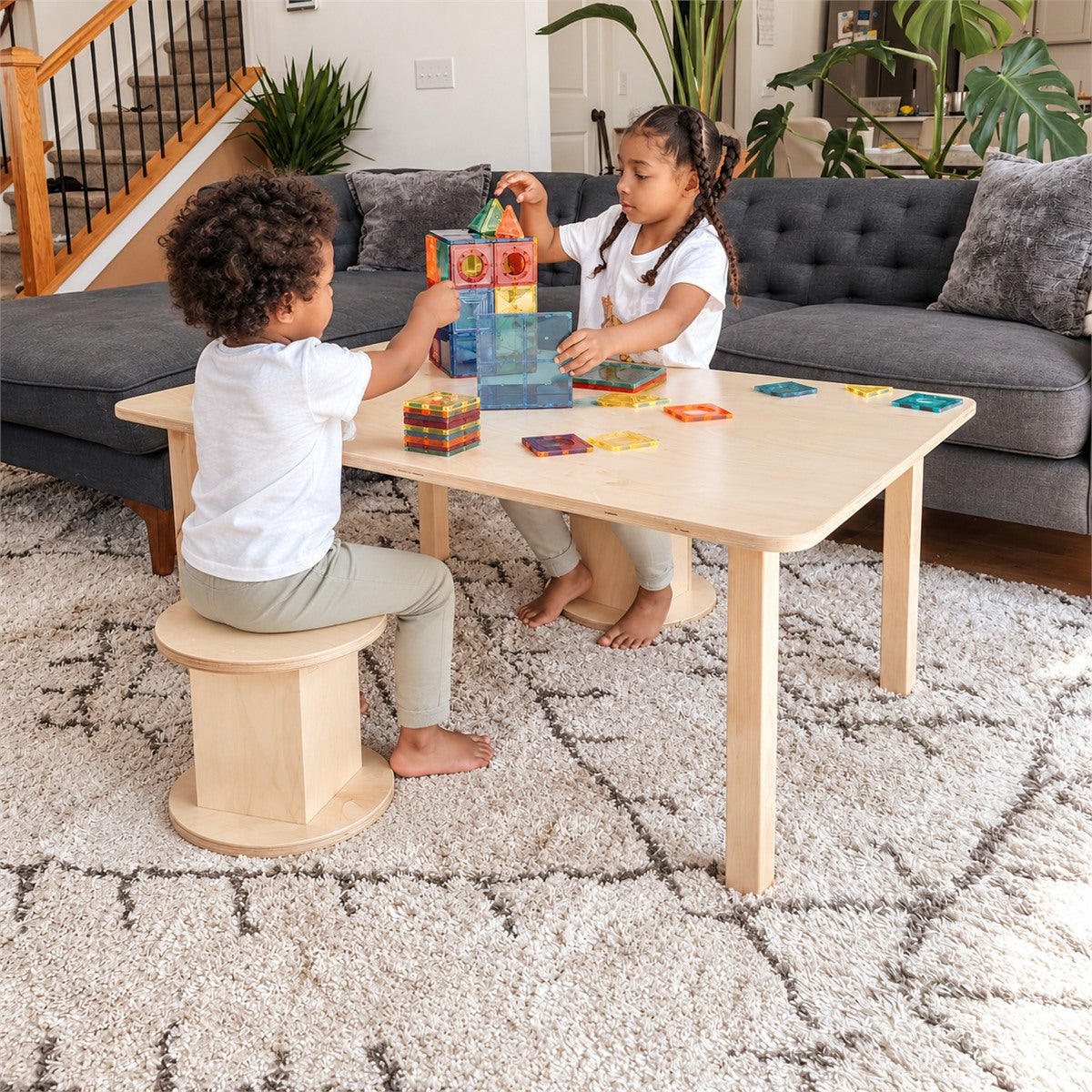 two children sitting at a wooden table with stools and magnatiles.
