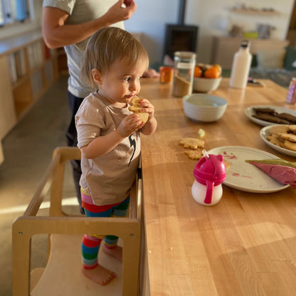Child standing on a chair at a dining table with food and a person in the background.