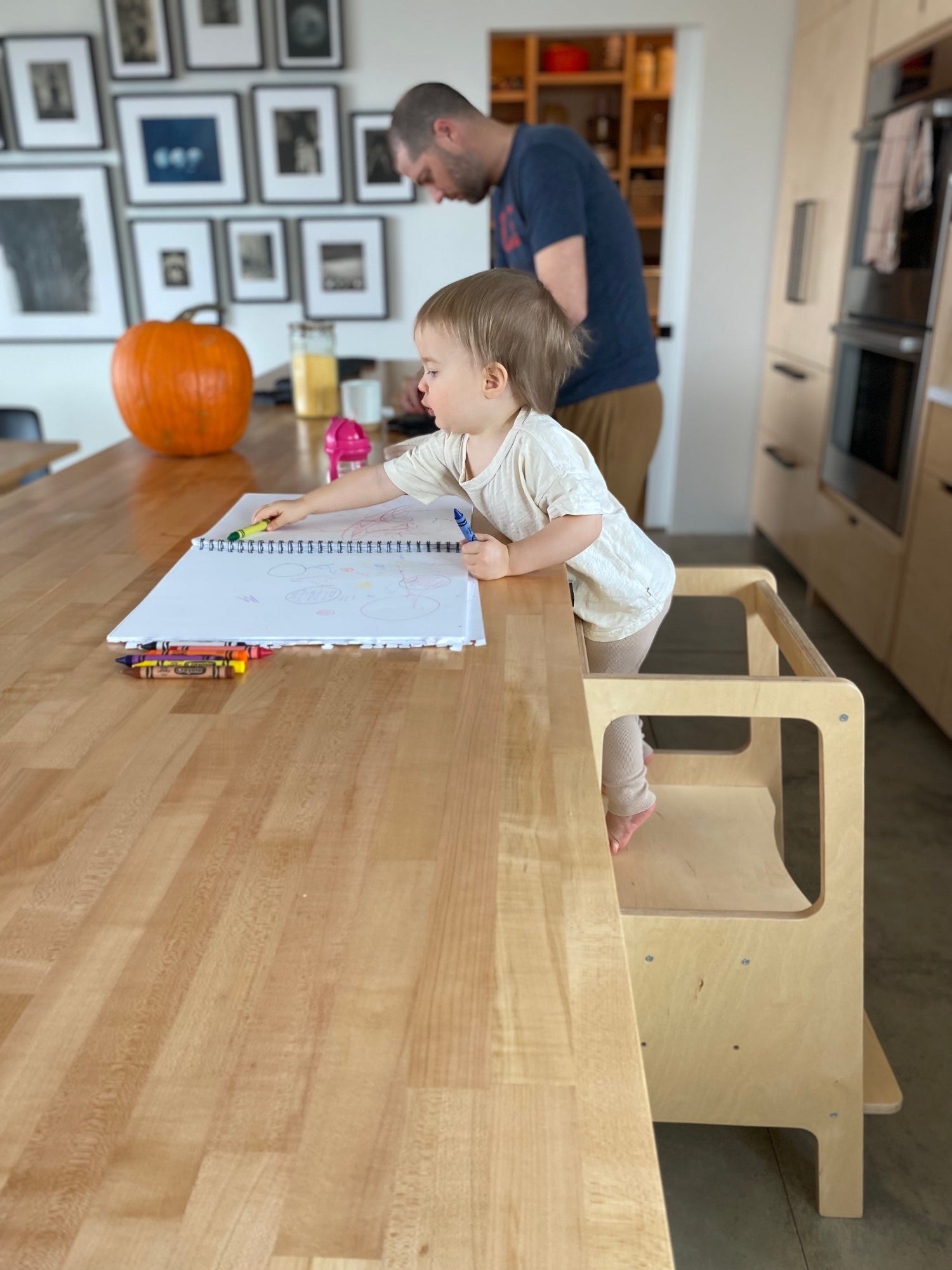 Child using a step stool to reach a table with a book and crayons, with an adult nearby.